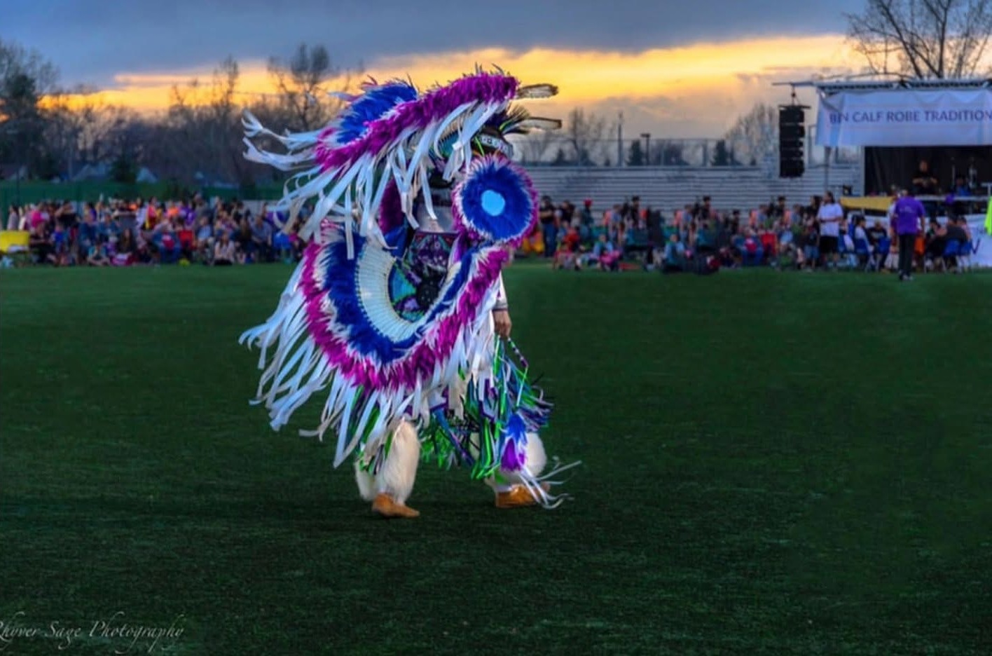 A fancy dancer in purple, blue and white regalia on the field at sunset, the Ben Calf Robe Tradition banner on the stage behind